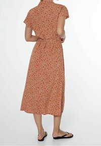 Rustinvärinen mekko, jossa on kukkakuosi, lyhyet hihat, kaulus ja vyötäröllä vyö. Keskipitkä, sivuhalkio ja kevyt kangas.