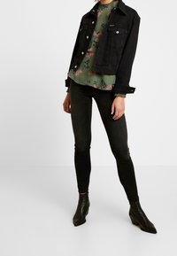 Person, der eine schwarze Denimjacke, eine grüne Bluse mit Blumenmuster, schwarze enge Jeans und schwarze spitze Stiefeletten trägt, steht vor einem weißen Hintergrund.