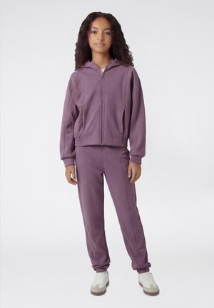 Joven de pie con una sudadera con capucha morada con cremallera y pantalones jogger a juego, y zapatillas blancas, sobre un fondo claro y liso.