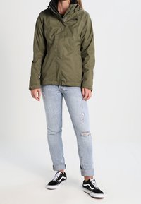 Grüne wasserdichte Jacke mit einer schwarzen Kapuze, getragen über einer hellblauen, zerrissenen Jeans und schwarz-weißen Turnschuhen.