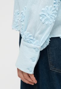 Μανίκι ανοιχτό μπλε μπλούζας με υφή, κεντημένο με λουλουδένια σχέδια και τρία κουμπιά, φορεμένο με σκούρο τζιν παντελόνι.