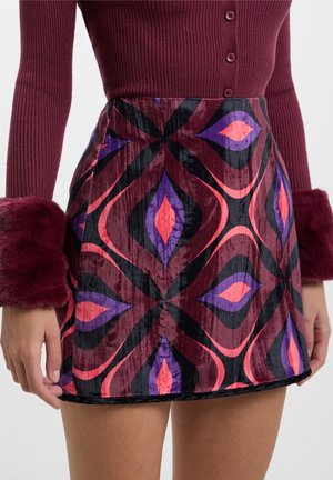 Frau trägt einen burgunderfarbenen gerippten Pullover mit pelzbesetzten Bündchen und einen hoch taillierten Samtrock mit lila, pinken und schwarzen geometrischen Mustern.