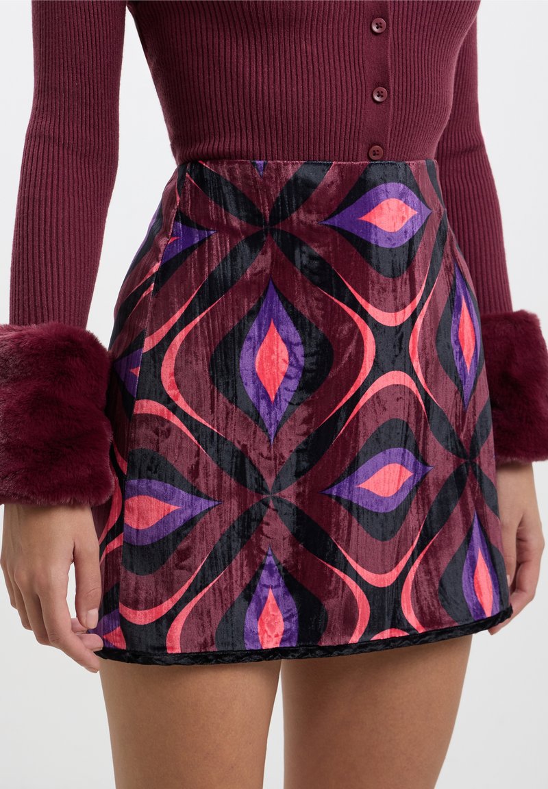 Femme portant un pull côtelé bordeaux avec des poignets en fourrure et une jupe en velours taille haute avec des motifs géométriques violet, rose et noir.