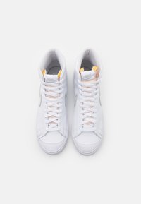 Bílé vysoké tenisky s koženým svrškem, polstrovaným límcem a tkaničkami. Obsahují šedé logo swoosh a žluté akcenty na jazyku.