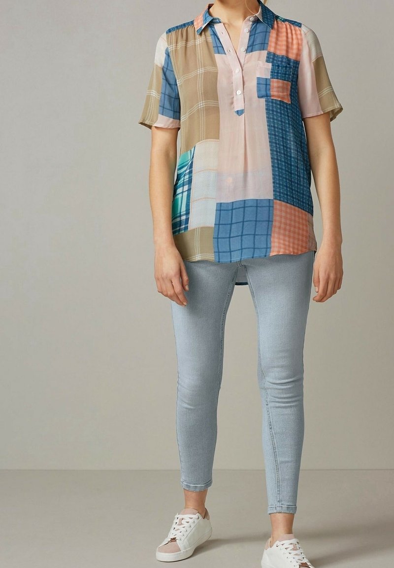 Femme portant une chemise patchwork multicolore à manches courtes, un jean skinny bleu clair et des baskets blanches, debout devant un fond uni.