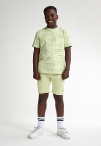 Lichtgroen T-shirt met korte mouwen en geometrische zwarte patronen, gecombineerd met bijpassende shorts. Witte sneakers en gestreepte sokken maken de look compleet.