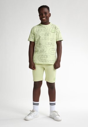 Jasnozielona koszulka z krótkim rękawem w geometryczne czarne wzory, zestawiona z dopasowanymi szortami. Białe sneakersy i paskowane skarpetki dopełniają całość.