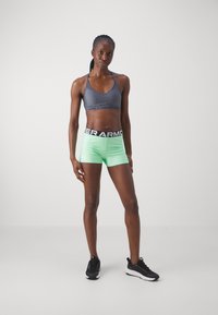 S sostenedor gris, pantalones cortos verdes de cintura alta con cinturilla "Under Armour" y zapatillas deportivas negras. Diseño minimalista, estilo ajustado.