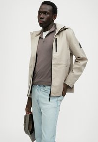 Bežna lahka jakna s kapuco, ki ima sprednji zadrgo in žepke z zadrgo, nosi pa se jo čez sivo pleteno majico in svetlo modre kavbojke.