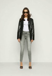 Jaqueta de couro preta sobre uma blusa branca com babados, combinada com jeans skinny cinzentos e sapatos de salto aberto pretos. Modelo usando óculos escuros escuros.