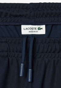 Ceinture élastique bleu marine avec cordons de serrage sur un vêtement, avec une étiquette blanche indiquant "LACOSTE REGULAR FIT" et un logo crocodile vert.