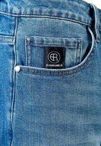 Šviesiai mėlynos spalvos džinsai su lygia tekstūra, turintys galinę kišenę su juodu etikete, kuriame matyti logotipas „ELIAS RUMELIS“.