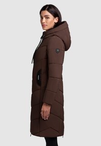 Palton pufos maro cu glugă, având un model cu diamante matlasat, buzunare laterale și un buton de accent negru pe mânecă.