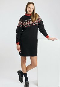 Robe tricotée noire avec un empiècement à motifs présentant des diamants rouges et blancs ; manches longues ; fentes latérales ; associée à des mocassins noirs et des chaussettes.