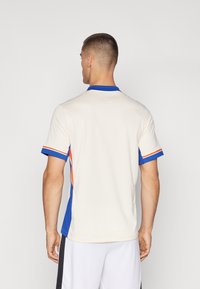 Herrtröja för atletisk användning med en krämfärgad kropp, blå krage och röda accenter. Korta ärmar med en strukturerad tyg. Visad bakvy.