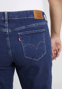 Tummansiniset farkut, joissa on nahkainen lappu, jossa lukee "Levi Strauss & Co." ja erottuva keltainen tikkauskuvio takataskuissa.