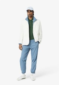 Blazer bianco, polo verde, giacca a vento azzurra chiara e pantaloni blu. Sneakers bianche con dettagli verdi, cappello a visiera piatta azzurro chiaro.