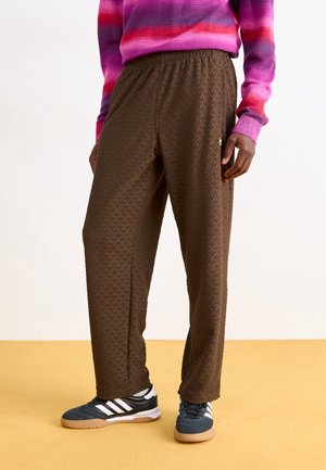 Personne portant un pantalon marron texturé, des baskets noires et blanches, et un haut à manches longues rayé rose, rouge et violet, debout sur un sol jaune.