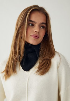 Jeune femme aux longs cheveux châtain clair, portant un pull blanc et une écharpe noire, regardant sur le côté devant un fond uni.