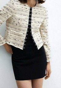 Veste en tweed couleur crème avec coutures texturées et accents noirs, dotée d'une fermeture à crochet. Robe noire cintrée en dessous.