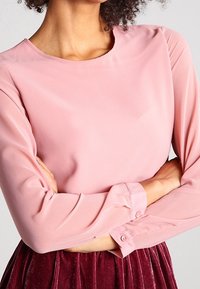 Rosa långärmad blus i mjukt tyg med rund halsringning, med knappade ärmslut. Mjuk struktur och kroppsnära design.