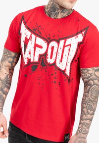 Rød bomulds T-shirt med et iøjnefaldende sort og hvidt "TAPOUT" logo med splattermønster, korte ærmer og et mærke ved kanten.
