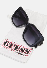 Ochelari de soare negri, oversize, cu lentile întunecate în degradé, de formă unghiulară. Însoțiți de un pachet alb cu un model de grilă roșie și logo-ul „GUESS” în negru.