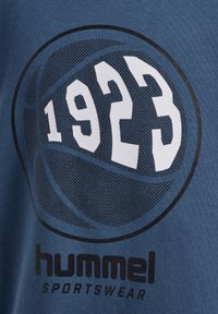 Niebieska koszulka z dużą czarno-białą grafiką koszykówki oraz napisem "1923" i "hummel SPORTSWEAR" poniżej w pogrubionej czcionce.