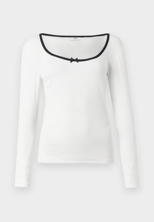 Langärmliges weißes Hemd mit quadratischem Ausschnitt, schwarzem Besatz und einem kleinen schwarzen Schleifendetail in der Mitte. Strukturierter, gerippter Stoff.