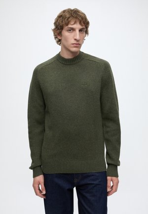 Jeune homme aux cheveux blonds ondulés porte un pull en laine vert olive et un jean bleu foncé, debout devant un fond clair uni.