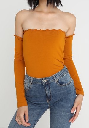 Femme portant un haut orange à épaules dénudées et manches longues, et un jean bleu taille haute, sur un fond clair uni.