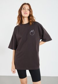 Brun oversized t-skjorte laget av bomull, med en enkel hvit linjetegning på brystet. Korte ermer og avslappet passform. Sorte sykkelshorts.
