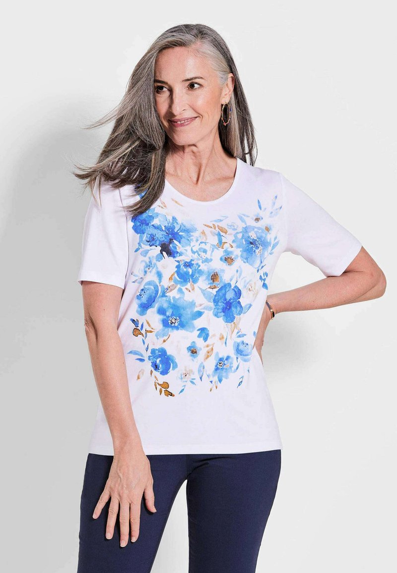 Witte T-shirt met korte mouwen, voorzien van een blauw bloemenpatroon met lichtbruine accenten aan de voorkant. Zachte stof met een ronde halslijn.