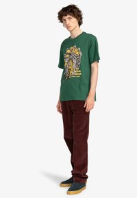 Camiseta de algodón verde con un estampado gráfico en amarillo y negro. Combinada con pantalones de pana burdeos y zapatillas verde oscuro.