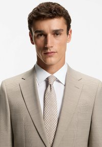Licht beige pak met gestructureerde stof, enkelrijig ontwerp, revers met inkeping, gecombineerd met een licht patroon das en wit overhemd.