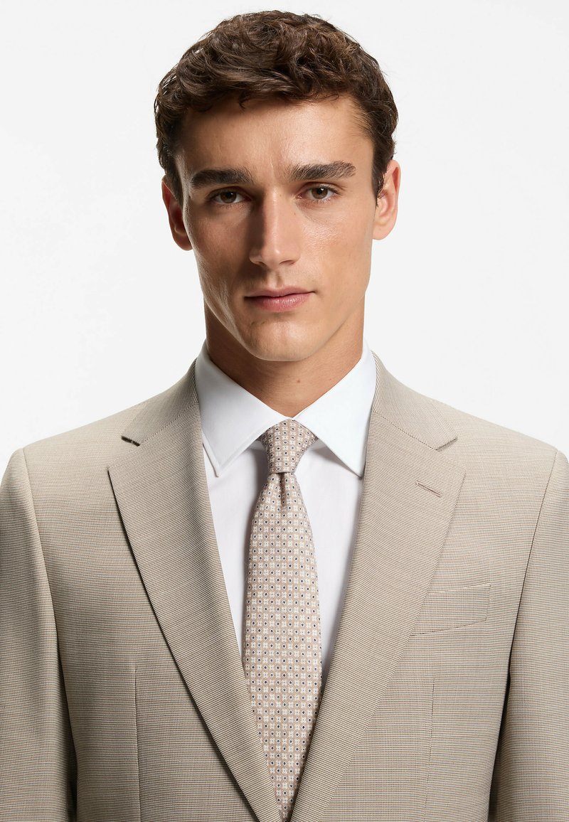 Licht beige pak met gestructureerde stof, enkelrijig ontwerp, revers met inkeping, gecombineerd met een licht patroon das en wit overhemd.