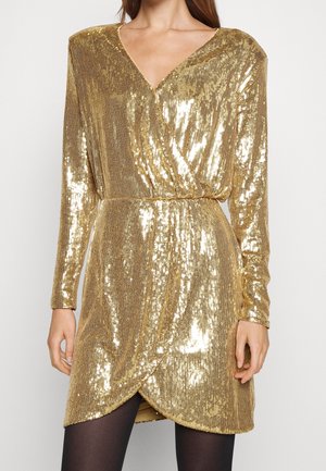 Femme portant une robe portefeuille à manches longues ornée de sequins dorés avec un col en V et des collants noirs sur un fond uni.