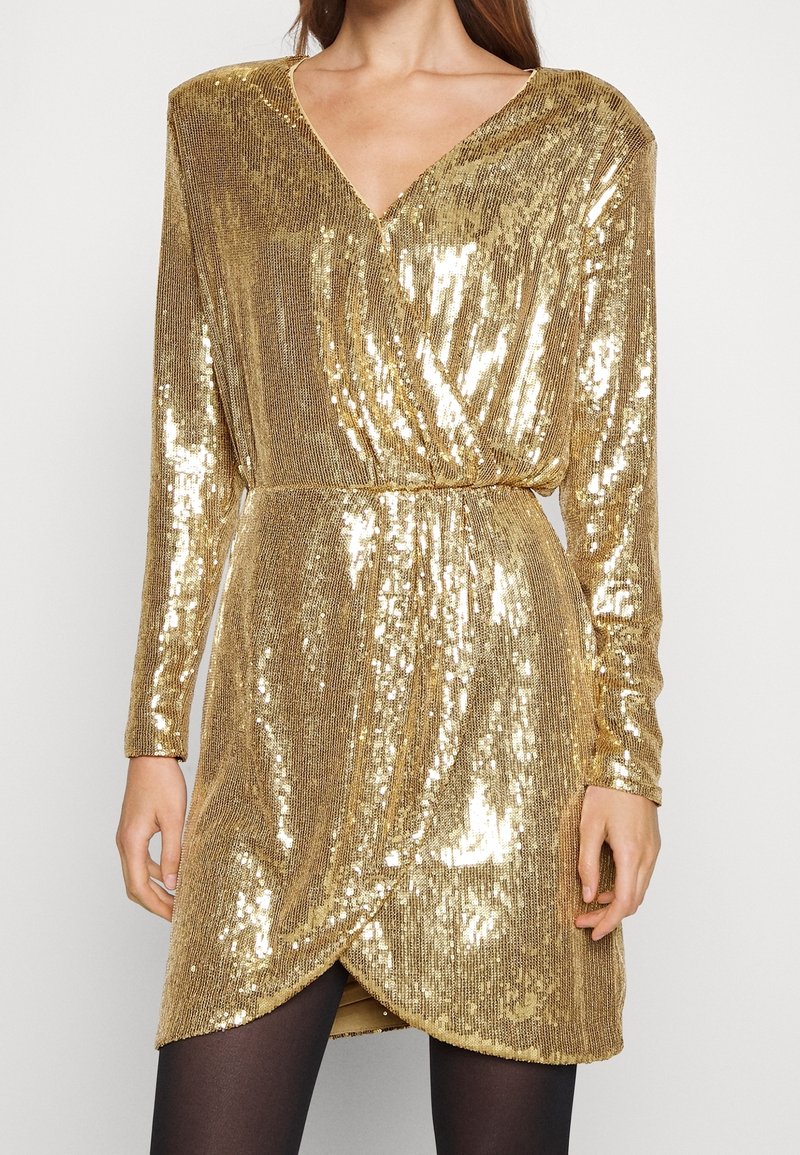 Femme portant une robe portefeuille à manches longues ornée de sequins dorés avec un col en V et des collants noirs sur un fond uni.