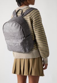 Gray leather backpack with a rounded shape, front pocket, and metal hardware details. Worn over a striped sweater and pleated skirt.