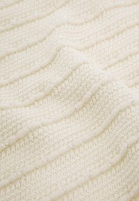 Couverture tricotée en blanc cassé avec motif ondulé. Présente une surface texturée et un léger éclat dû aux accents tissés. Tissu doux et confortable.