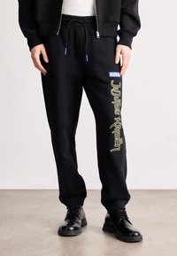 Μαύρες φόρμες προσφοράς με κορδόνι στη μέση, πλάγιες τσέπες και εντυπωσιακό κίτρινο κείμενο "Jujutsu Kaisen", καθώς και μπλε ετικέτα "HUGO".