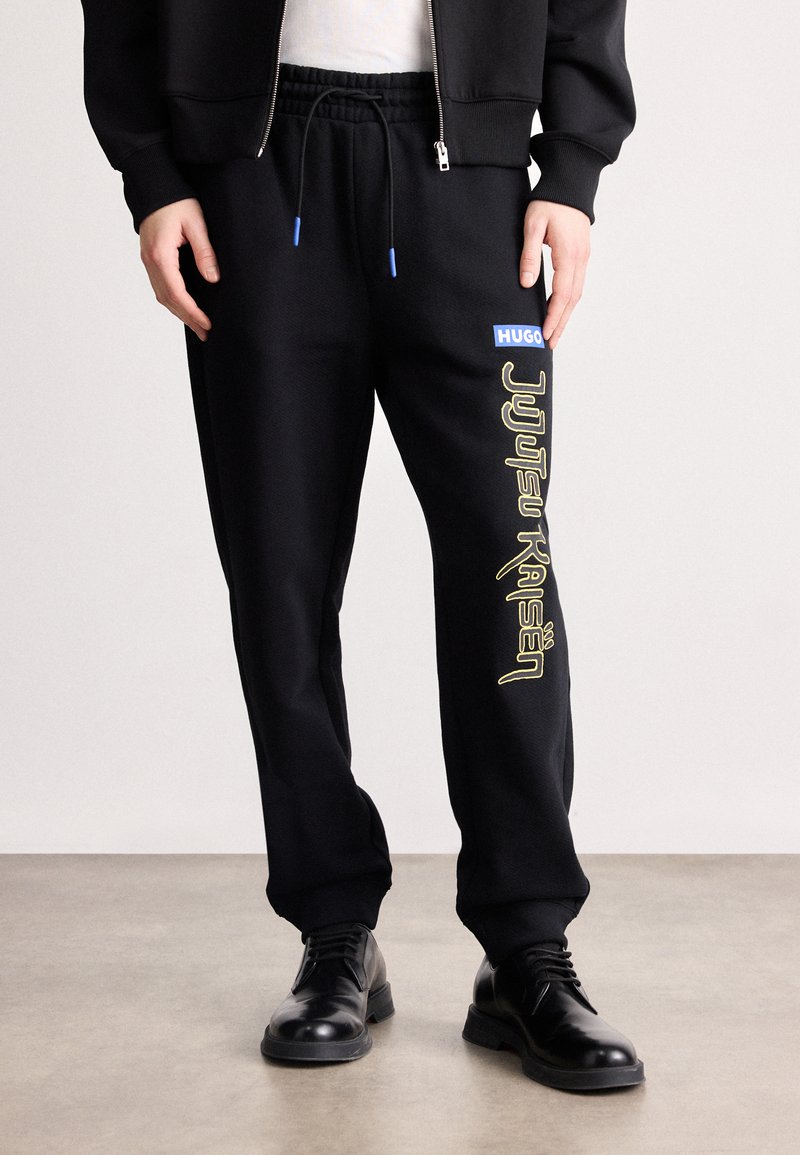 Μαύρες φόρμες προσφοράς με κορδόνι στη μέση, πλάγιες τσέπες και εντυπωσιακό κίτρινο κείμενο "Jujutsu Kaisen", καθώς και μπλε ετικέτα "HUGO".