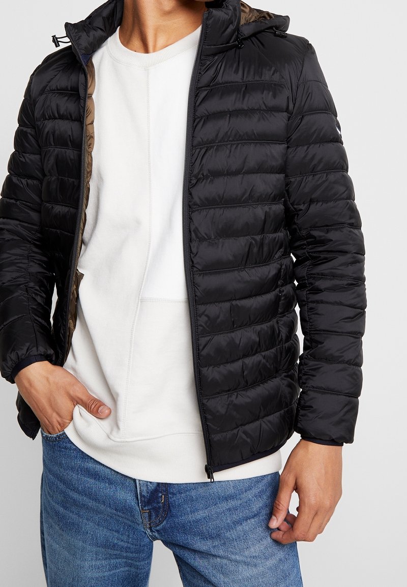 Schwarze Pufferjacke mit horizontalen Nähten, Reißverschluss vorne und verstellbarem Kapuzenkragen. Getragen über einem weißen Sweatshirt und blauen Jeans.