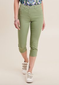 Jeans capri verdi con un design aderente, realizzati in tessuto elastico, dotati di tasche anteriori e una superficie liscia, abbinati a scarpe bianche.