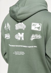 Grüne Kapuzenjacke mit verschiedenen weißen Logos und Texten. Das Material wirkt weich; sie hat eine Kapuze und ein bedrucktes Design auf der Rückseite.