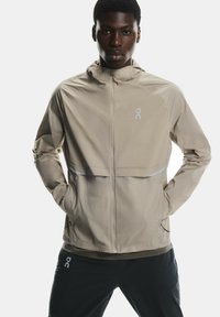 Viegls, beige skrējēju džemperis ar rāvējslēdzēju priekšā, sānu kabatām un atstarojošiem akcentiem. Izgatavots no gludas, ūdensizturīgas auduma ar kapuci.