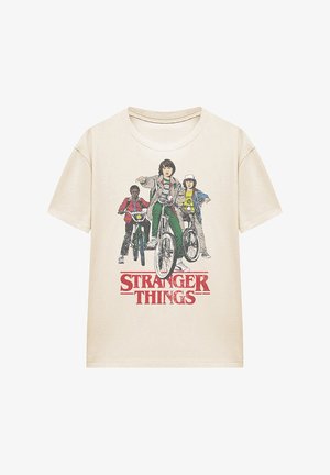 Beige cotton t-shirt featuring a graphic print of three characters on bicycles, with "STRANGER THINGS" text in red underneath.