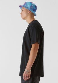 T-shirt en coton noir à manches courtes, associé à un chapeau seau tie-dye bleu et violet. Le modèle se tient de côté, exhibant des tatouages et des bijoux.