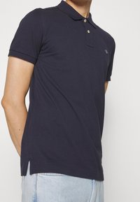 Marinblå polo skjorta i bomull med korta ärmar, krage och två-knapps placket. Har sidslitsar och en liten broderad logotyp.