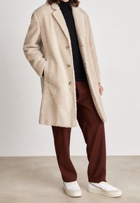 Persona che indossa un cappotto beige a trama, dolcevita nero, pantaloni marroni e sneakers bianche, in piedi su un pavimento chiaro contro un muro semplice.